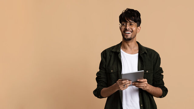 A smiling young man stands confidently holding a tablet in his hands, dressed casually. The plain beige background adds warmth to the scene, highlighting his cheerful expression.