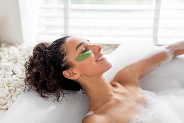 Naklejka premium A woman enjoys a tranquil moment soaking in a bubble-filled bath while wearing a green face mask. Soft sunlight filters through a window, enhancing the peaceful atmosphere.