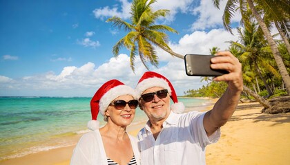 Happy senior couple in Santa hats taking a selfie on a bright tropical beach with palm trees. Fantastic holidays destination with clear sky and turquoise water.