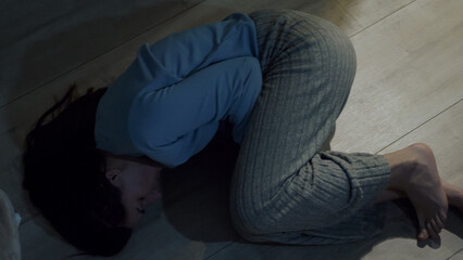 A young woman lies curled on the floor in a fetal position, holding herself tightly. A moment of emotional collapse, deep vulnerability, and inner struggle in a dramatic low-light setting. 