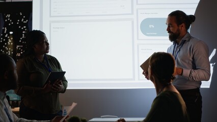 Multiethnic team of workers discuss the financial situation of the company, presenting data forecasts and diagrams to advisory board. Attend a meeting late at night in the office. Camera B.