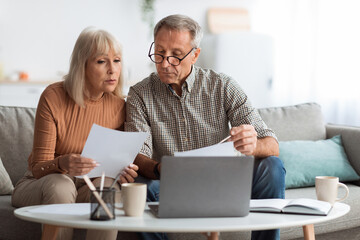 An older couple sits closely on a sofa reviewing important documents. They appear focused and engaged in a discussion. A laptop is open in front of them, adding to the cozy atmosphere.