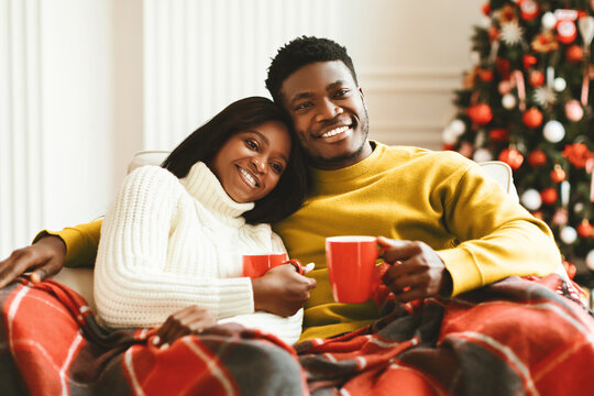 A couple sits together on a sofa holding red mugs filled with warm drinks. They are wrapped in cozy blankets and are surrounded by a decorated Christmas tree. The setting is festive. - Powered by Adobe
