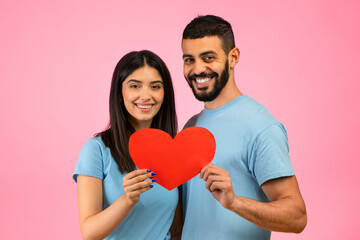 A young couple smiles widely while holding a large red heart in front of a bright pink background. They are dressed in matching light blue shirts, showcasing love and happiness.