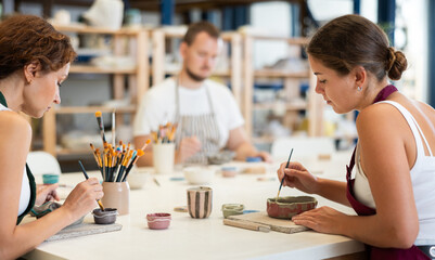In pottery workshop, two girls and guy are sitting at table, busy coloring products made of raw clay. Amateurs are busy painting clay plates at master class