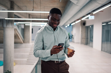 African american businessman using phone holding coffee in modern office