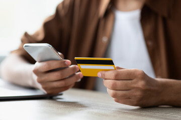 Individual engages in online shopping, holding a smartphone in one hand and a yellow credit card in...