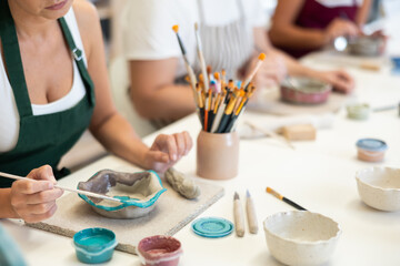 Close-up of girl employee, she decorates and paints clay product, works in company of craftsmen and amateurs. Workshop for manufacture of authors products