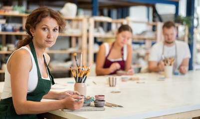 During working day, three employees of pottery workshop paint finished product from raw clay. They apply pattern to surface of plate with brush, dye decorative plate, carry out order at work table