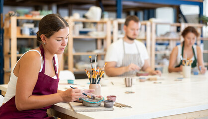 During working day, three employees of pottery workshop paint finished product from raw clay. They apply pattern to surface of plate with brush, dye decorative plate, carry out order at work table