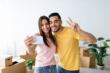 A couple smiles and poses for a selfie in their new home filled with moving boxes. They are happy and excited about their new beginning.