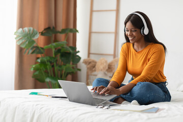 A woman sits cross-legged on her bed, smiling as she types on her laptop. She wears headphones and...