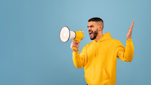 A man in a bright yellow hoodie is energetically shouting into a megaphone, expressing strong emotions. The background is a solid light blue, highlighting his actions and passion. - Powered by Adobe
