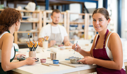In pottery workshop, two girls and guy are sitting at table, busy coloring products made of raw clay. Amateurs are busy painting clay plates at master class