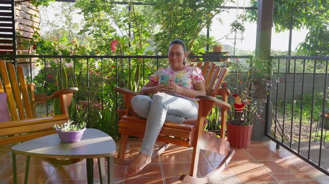 Latina woman smiling at smartphone while relaxing barefoot on sunny porch