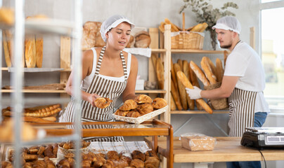 Smiling young adult female worker puts croissants in the case as her colleague stacks baguettes