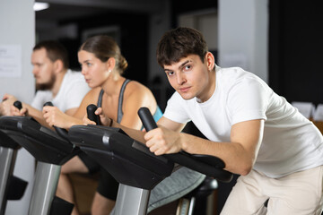 In gym, guy does exercises using stationary bicycle. Young group of athletes trains in gym. Physical education and sports, active healthy lifestyle.