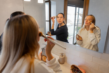 Two friends in modern home bathroom applying face powder with brushes while looking in mirror. Beauty, daily routine, cosmetics and bonding. Indoor horizontal close-up with natural light from balcony.