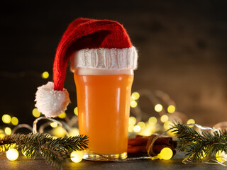 Craft Beer glass in a knitted Christmas hat on a table with Christmas tree branches and a bright garland, copy space