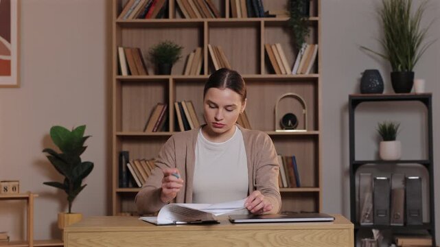 Caucasian woman sitting at wooden desk reading papers and marking notes. Female concentrating on work reviewing documents at home workspace with laptop and organized shelves filled with books behind.