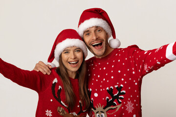 A happy couple poses together, wearing red Christmas sweaters with playful designs and Santa hats. They are smiling joyfully, embracing the festive spirit of the season.