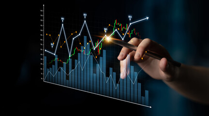 A hand holds a pen as it analyzes stock market trends represented by graphs and charts on a digital...