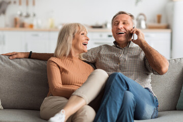 A cheerful couple sits closely on a couch, laughing together. The man is on the phone while the woman smiles, creating a warm and lively atmosphere in their home.