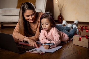 A mother and her young daughter are lying on the floor, engaging in a drawing activity. They are surrounded by gifts and a laptop, creating a warm atmosphere in their home.