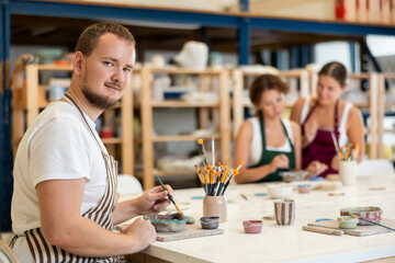 Male apprentice, is sitting near desktop and coloring clay product. In background, lovers of applied art complete design of bowl made of clay, apply pattern with brush in presence of teacher
