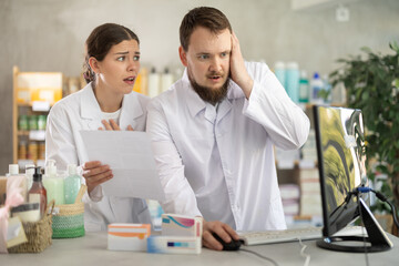 Confused male pharmacist and female assistant discuss financial problems of pharmacy. Employees use paper and electronic documents to find source of financial problems.