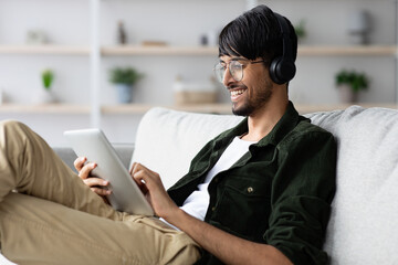 A young man with glasses and headphones smiles while using a tablet. He is lounging on a light-colored couch in a bright living room with plants in the background.