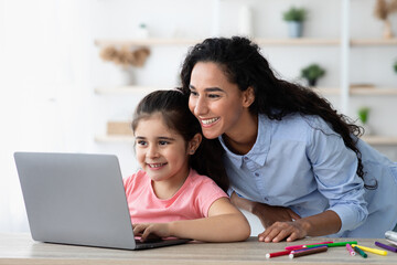 A mother and daughter share a joyful experience as they look at a laptop screen. Situated in a bright room, they are engaged in an educational activity. Colorful pencils are scattered nearby.