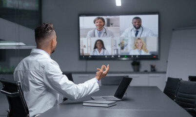 A group of medical professionals are engaged in a video conference. One person speaks while others listen and participate. The setting is a modern conference room with a large screen.