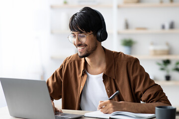 A young man is engaged in online learning at home, wearing headphones and smiling while taking notes. His workspace is simple and well-lit, featuring a laptop and a notebook.