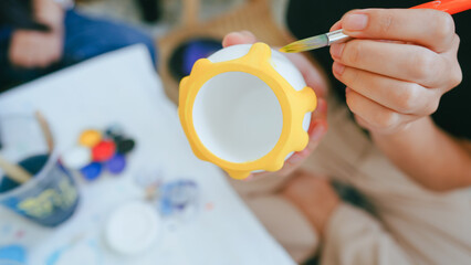 Close-up of a person hand painting a ceramic pot with yellow acrylic paint during an art workshop or creative DIY home project.