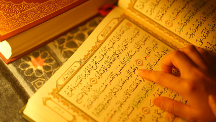 Muslim hand pointing at verse in Quran with warm lighting, focusing on Arabic script and Islamic holy book during worship or spiritual study moment.