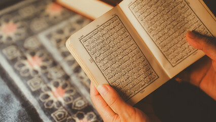 Close-up of hands holding and reading the Quran with Arabic script during spiritual prayer time on a patterned prayer rug.