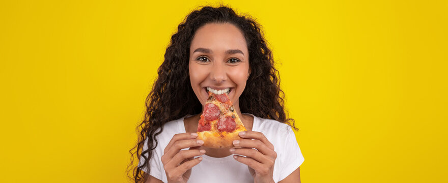 A young woman with curly hair is smiling while holding a slice of pizza. The vibrant yellow background enhances her joyful expression as she prepares to take a bite. - Powered by Adobe