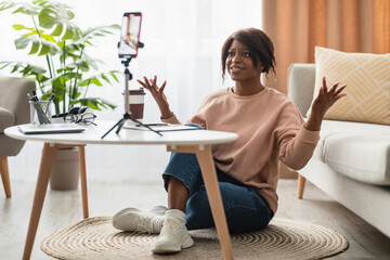 A woman dressed in casual attire sits on the floor in her cozy living room. She gestures animatedly while speaking to a smartphone mounted on a tripod. The atmosphere is bright and inviting.