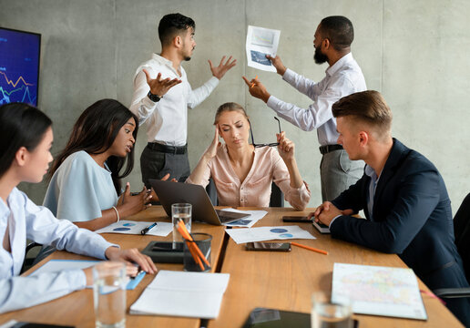In a bright, modern boardroom, a group of stressed business people are having a disagreement. Two male coworkers passionately discuss a financial report while others listen intently.