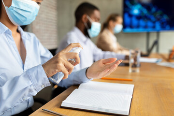 In a modern office, a female employee wears a mask and sprays disinfectant on her hands while attending a corporate meeting with diverse coworkers.