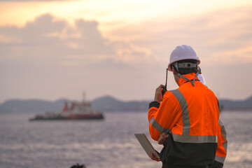 Two engineers wearing safety uniforms and hard hats communicate using a radio while reviewing data on a laptop by the sea during sunset. Maritime industrial setting.
