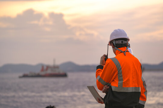 Two engineers wearing safety uniforms and hard hats communicate using a radio while reviewing data on a laptop by the sea during sunset. Maritime industrial setting.
