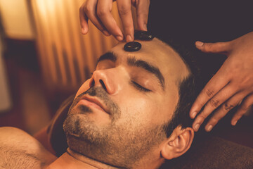 Fototapeta premium Caucasian man enjoying relaxing anti-stress head massage with hot stone and pampering facial beauty skin recreation leisure in warm candle light ambient salon spa in resort or hotel. Quiescent