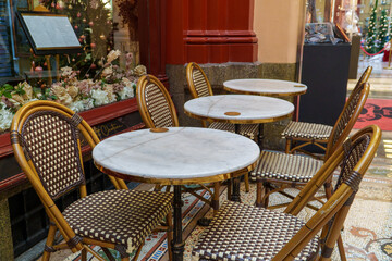 Charming outdoor cafe seating with round marble tables and rattan chairs in a festive, decorated arcade setting.