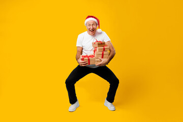 A man stands against a yellow background with multiple gift boxes in his hands. He wears a Santa hat and has a big smile on his face, celebrating the holiday spirit.