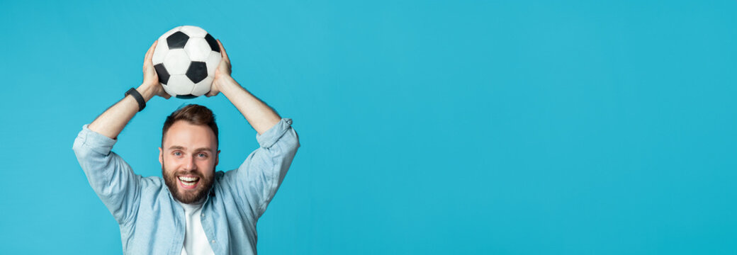 A joyful man with a beard is smiling and raising a black and white soccer ball above his head. He is wearing a light blue shirt and shows his excitement in a bright blue setting. - Powered by Adobe