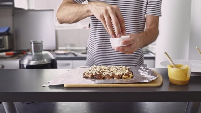 A person carefully adds chopped nuts to a delicious chocolate dessert, freshly prepared on a kitchen counter. The homemade treat promises a sweet and crunchy experience for any occasion.