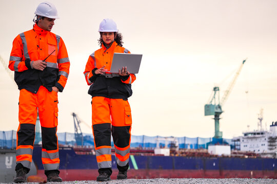 Two port engineers in high-visibility orange uniforms and hard hats communicate while inspecting dockside logistics using a laptop and walkie-talkie, symbolizing teamwork and safety.