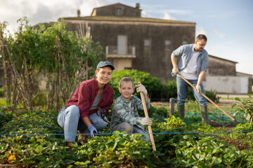 Fototapeta premium Woman farmer with her son and garden tools after planting strawberries. Family of farmers growing organic vegetables in the garden